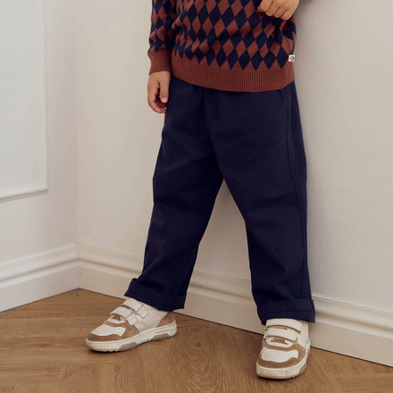 Child wearing a patterned sweater and navy pants standing against a white wall.
