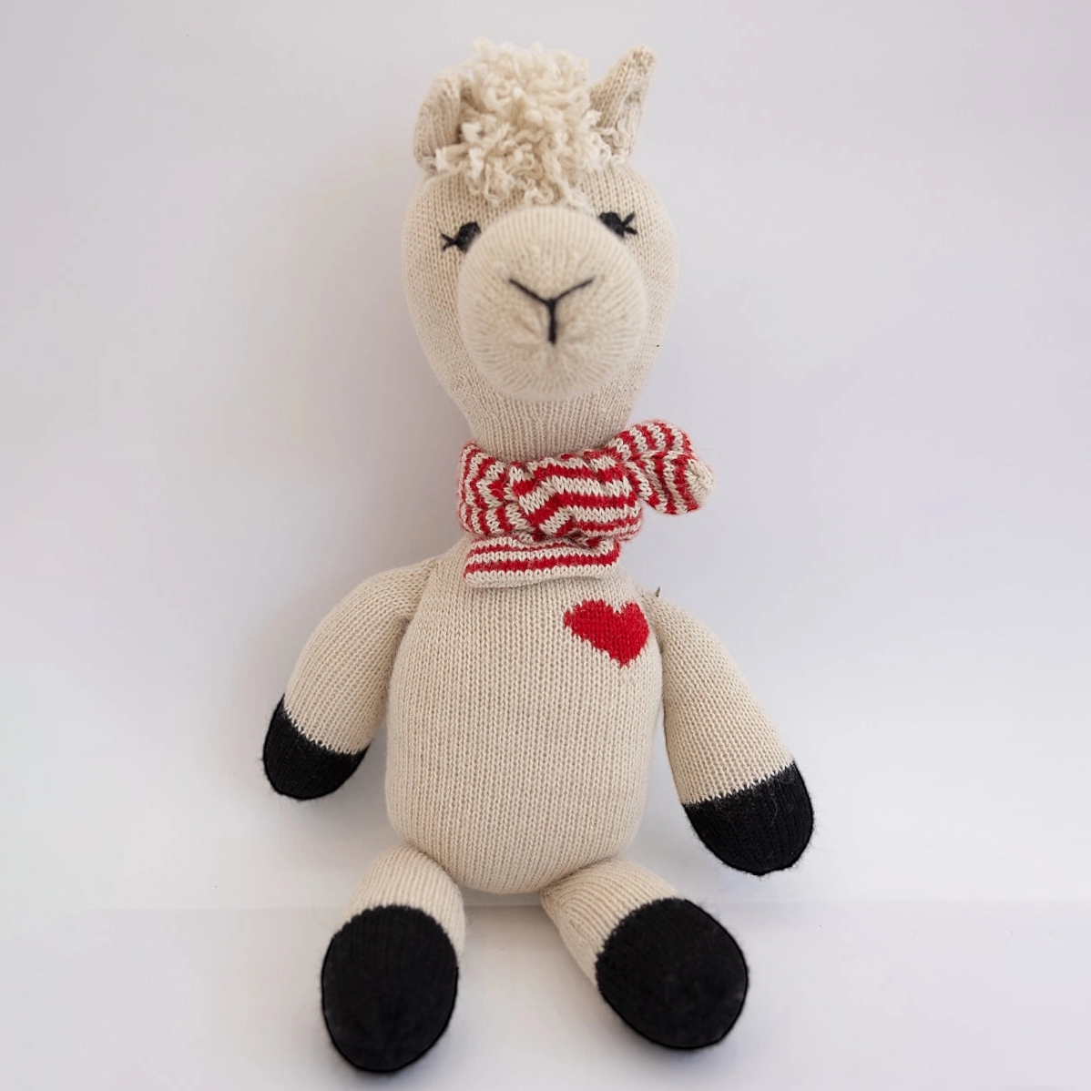 Knitted alpaca toy with a red heart and striped scarf on a white background