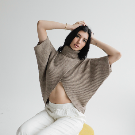 Woman wearing a beige knitted poncho over white pants on a light gray background