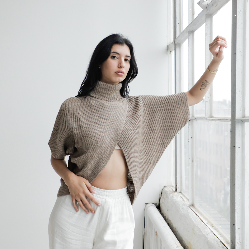 Woman wearing a beige knitted poncho in a bright room with a window.