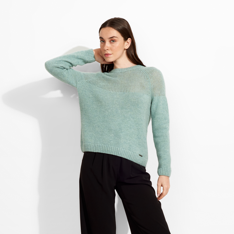 Woman wearing a light green sweater and black pants against a white background