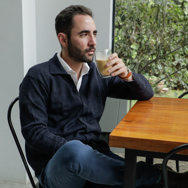 Man sitting at a wooden table holding a coffee, with a blurred outdoor background.