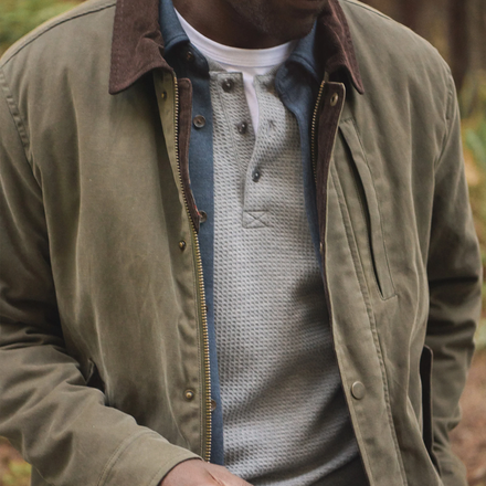 Person wearing a green jacket with a brown collar and a gray sweater underneath, standing outdoors.