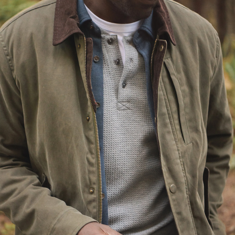 Person wearing a green jacket with a brown collar and a gray sweater underneath, standing outdoors.