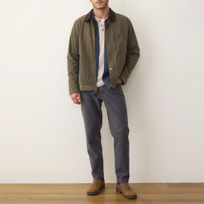 Man wearing a green jacket, blue shirt, gray pants, and brown shoes against a white wall.