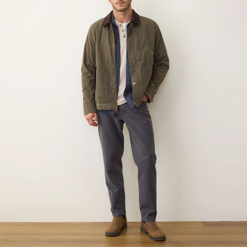 Man wearing a green jacket, blue shirt, gray pants, and brown shoes against a white wall.