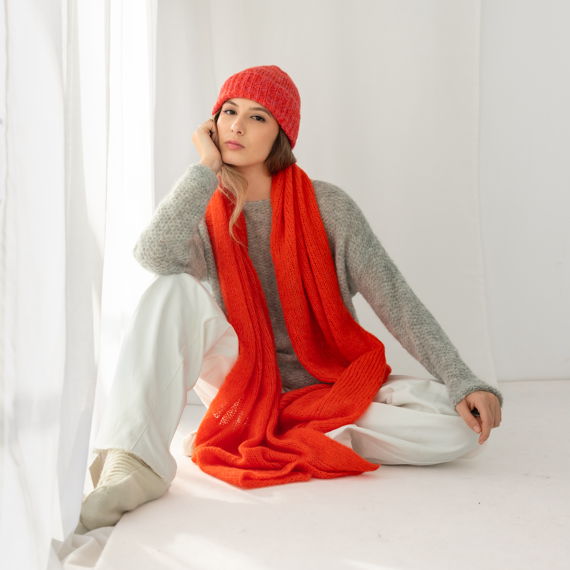 Person wearing a red shawl and hat sitting on a white surface with a white curtain in the background