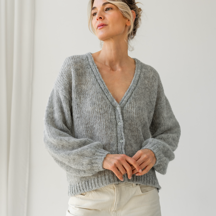 Woman wearing a gray knit cardigan and white pants against a plain background