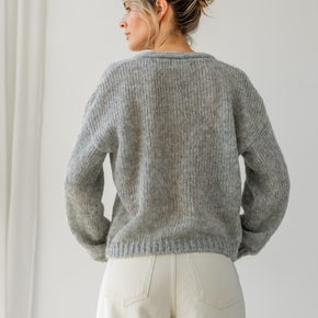 Woman wearing a gray knitted sweater and white pants against a plain background