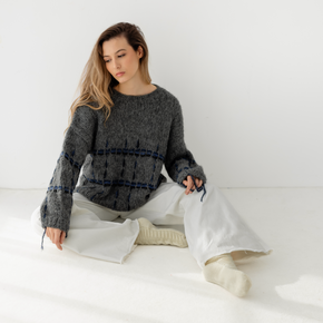 Woman sitting on a white floor wearing a dark gray sweater with blue and black accents and light pants.