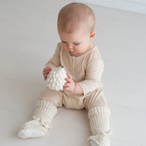 Baby in a beige outfit sitting on a light-colored floor holding a white toy.
