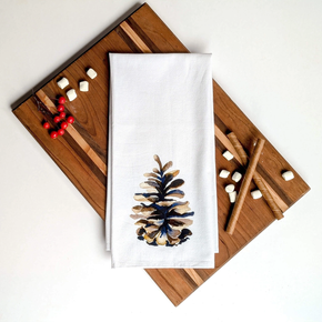 White towel with pine cone design on a wooden cutting board with cinnamon sticks and berries.