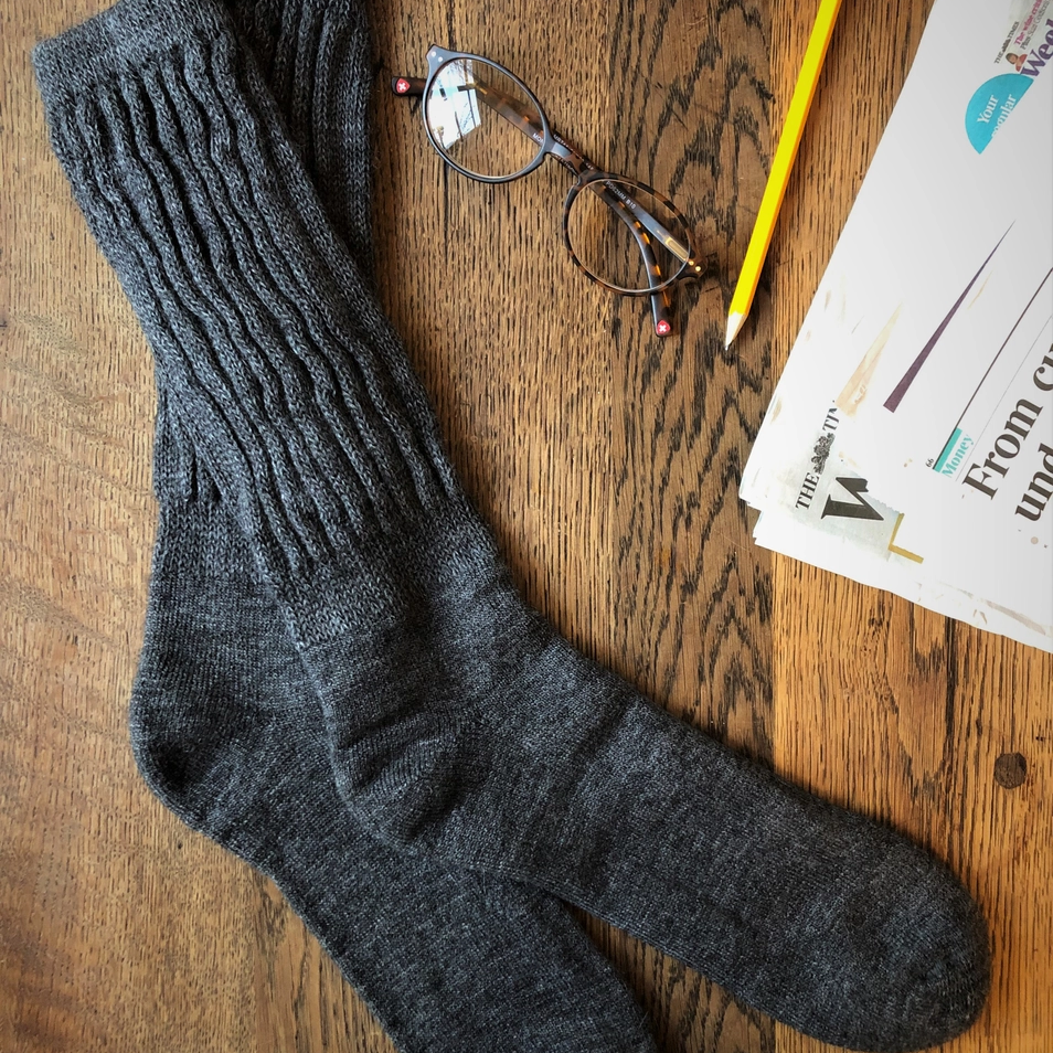 A pair of rib knit alpaca socks in gray, displayed on a wooden surface, accompanied by a pair of glasses and a pencil, with part of a newspaper visible in the background.