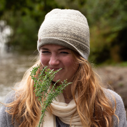 Woman wearing a knit hat and scarf holding a plant by a body of water.