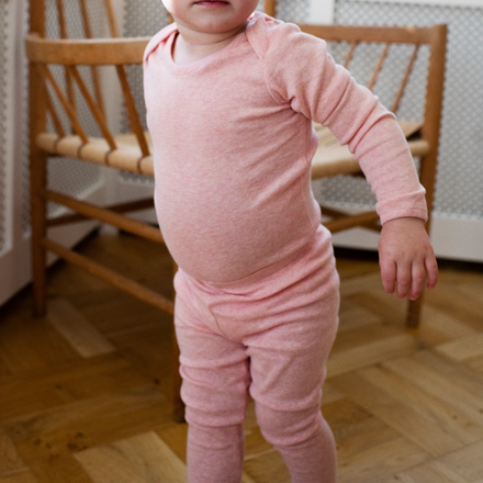 Child wearing a pink outfit standing on a wooden floor with a chair in the background.