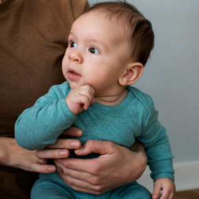 Baby in a teal onesie being held by an adult on a brown couch.