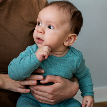Baby in a teal onesie being held by an adult on a brown couch.