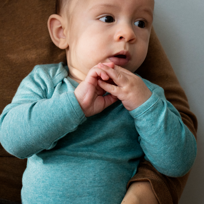 Baby in a teal onesie sitting on a person's lap with a neutral background