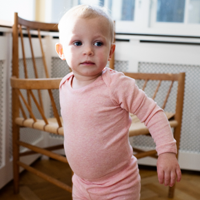 Child wearing a pink outfit standing on a wooden floor with a chair in the background.