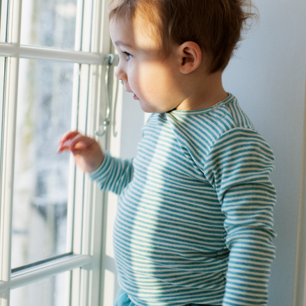 Child wearing a teal and white striped shirt  standing by a window.