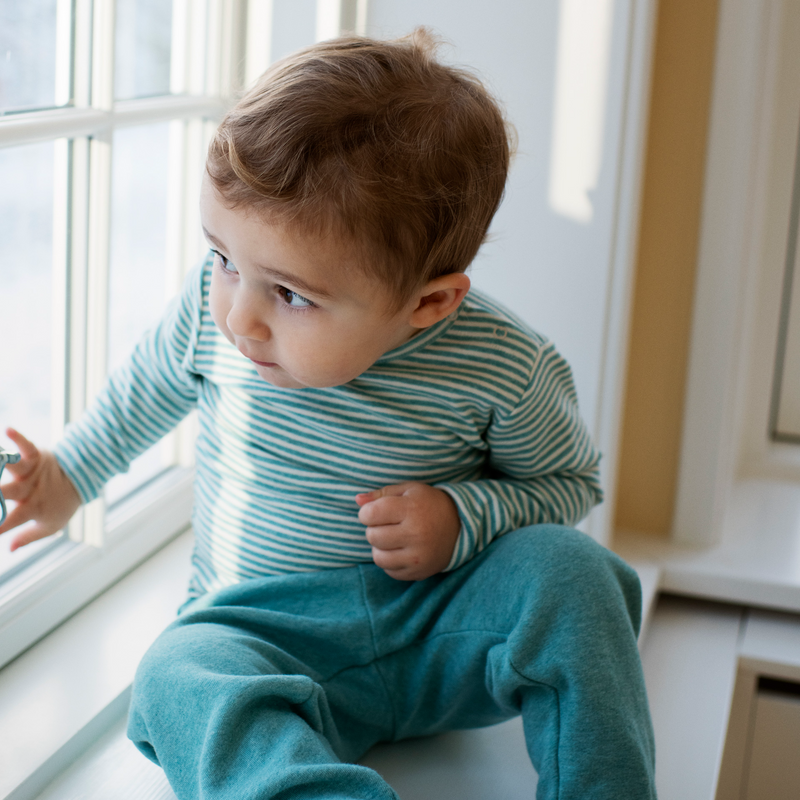Child wearing a teal and white striped outfit sitting by a window.