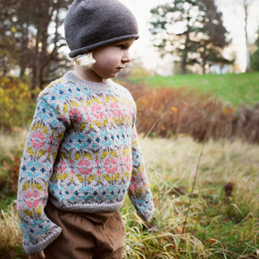 Child wearing a colorful sweater and gray beanie standing in a natural setting with trees and grass.
