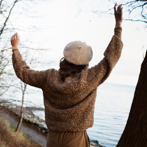 Person wearing a brown sweater and beige beret with arms raised by a body of water.