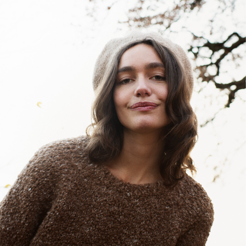 Woman wearing a brown sweater and beige beret standing outdoors with trees in the background