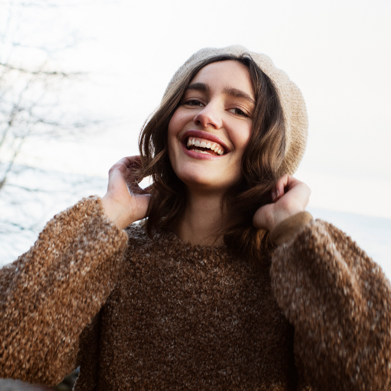 Woman wearing a brown sweater and beige beret outdoors with trees in the background