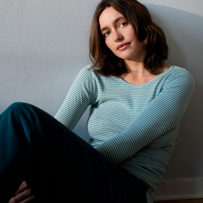 Woman wearing a light blue striped shirt sitting against a plain wall.