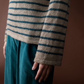 Person wearing a striped sweater and teal pants against a brown background