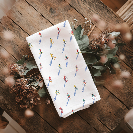 White tea towel with colorful skier pattern on a wooden table with dried flowers.