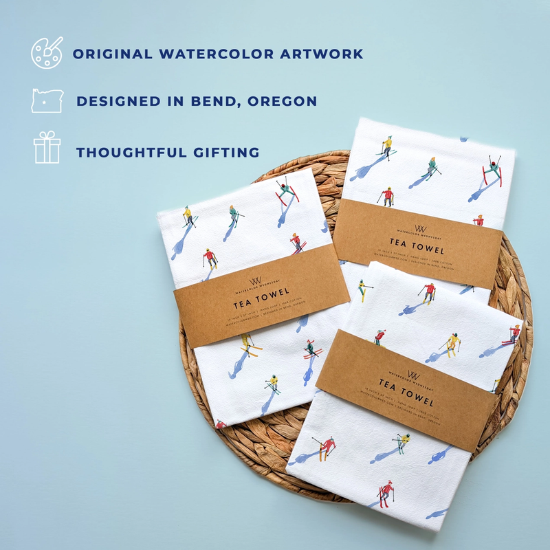Set of tea towels with artistic packaging on a woven tray against a light blue background.