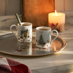 Two mugs with Christmas designs on a tray with a lit candle in the background.