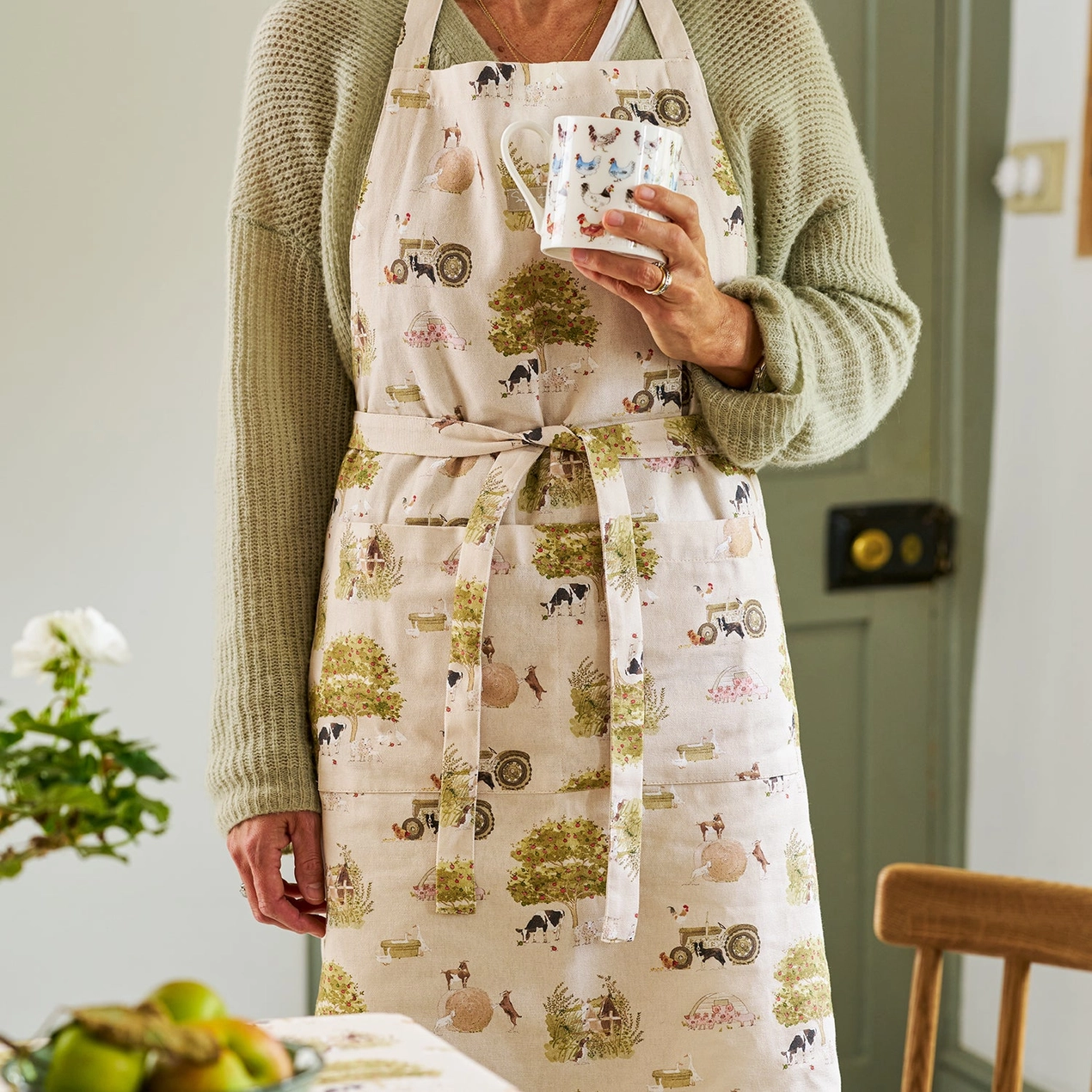 Person wearing a patterned apron holding a mug in a kitchen setting.