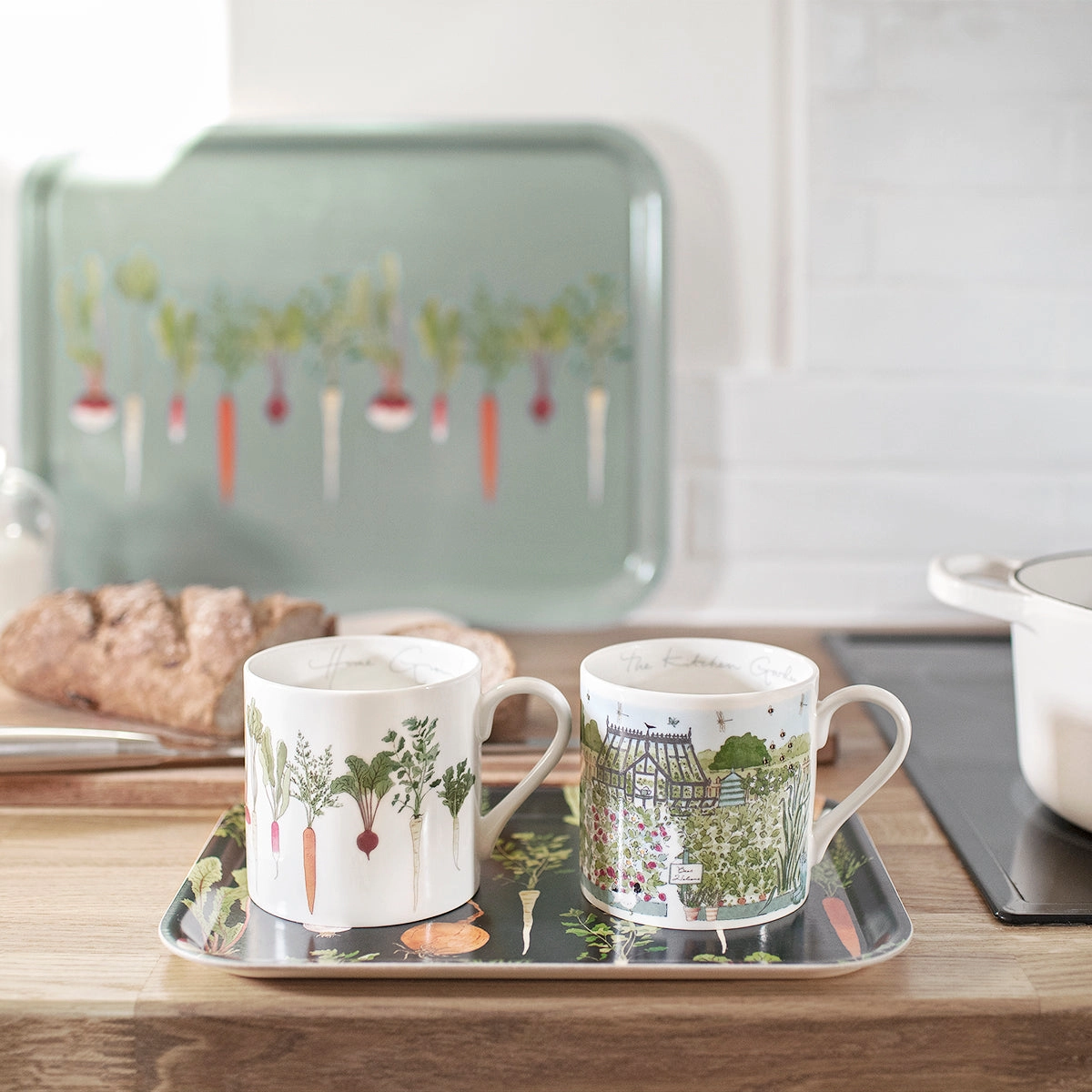 Two mugs with garden-themed designs on a tray with bread and a teapot in the background.