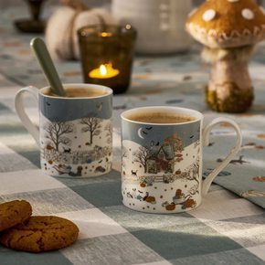 Two mugs with autumn-themed designs on a checkered tablecloth with cookies and a candle.
