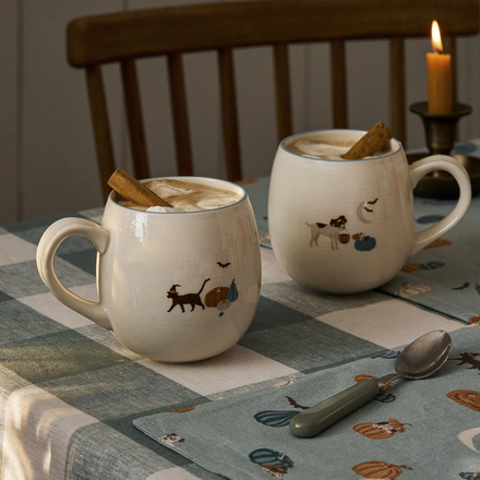 Two ceramic mugs with animal illustrations on a checkered tablecloth with a candle in the background.
