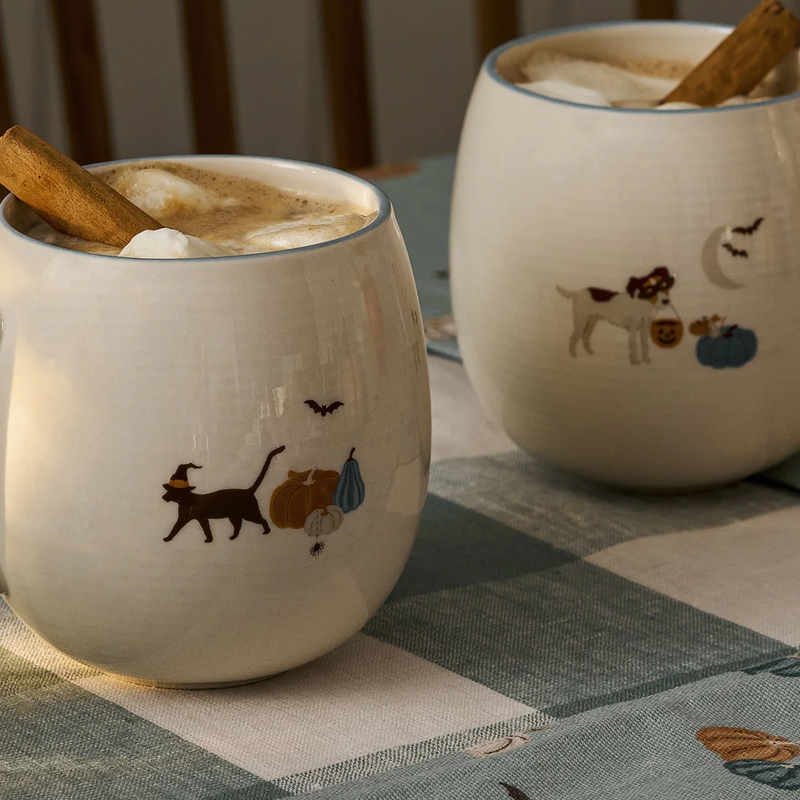 Two ceramic mugs with Halloween-themed illustrations on a checkered tablecloth.