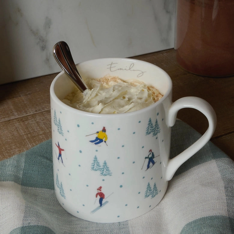 White mug with skiing patterns filled with a hot beverage and whipped cream, on a checkered cloth.