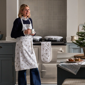 Woman in a kitchen holding a cup, wearing an apron and oven mitt, with a Christmas tree in the background.