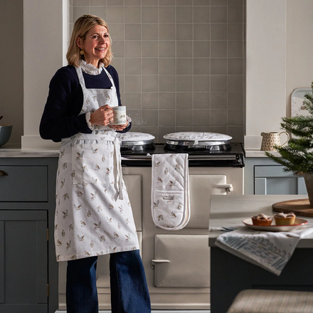 Woman in a kitchen holding a cup, wearing an apron and oven mitt, with a Christmas tree in the background.