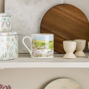 A white fine bone china mug with a spring cottage scene design, including a cottage, blossoming trees, and baby animals.