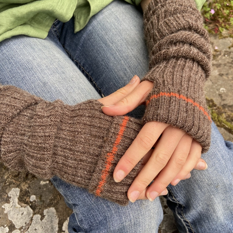 Close-up of brown knitted arm warmers with orange accents worn by a person sitting on a stone surface.
