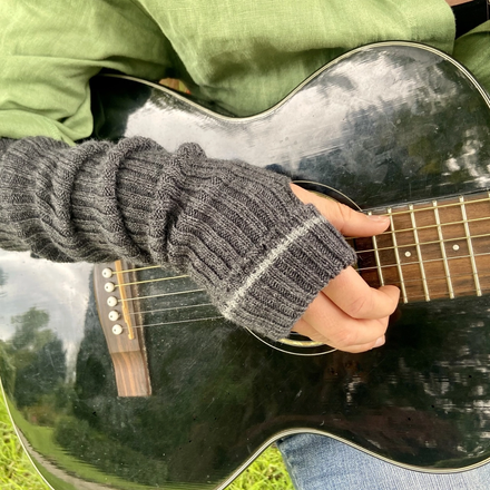 Person playing an acoustic guitar with a blurred natural background, wearing grey wrist warmers with lighter grey stripe
