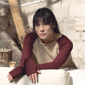 Woman wearing a burgundy long-sleeve top over a beige sweater against a stone wall.
