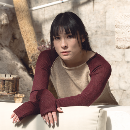 Woman wearing a burgundy long-sleeve top over a beige sweater against a stone wall.