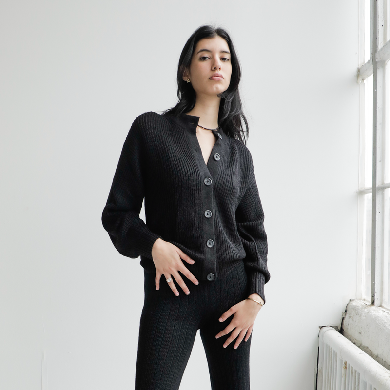Woman wearing a black cardigan standing in a room with a white wall and window.