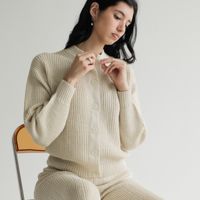 Woman wearing a cream knit outfit sitting on a chair against a plain background
