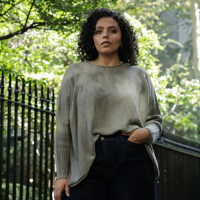 Woman wearing a green sweater and dark pants standing outdoors with trees in the background